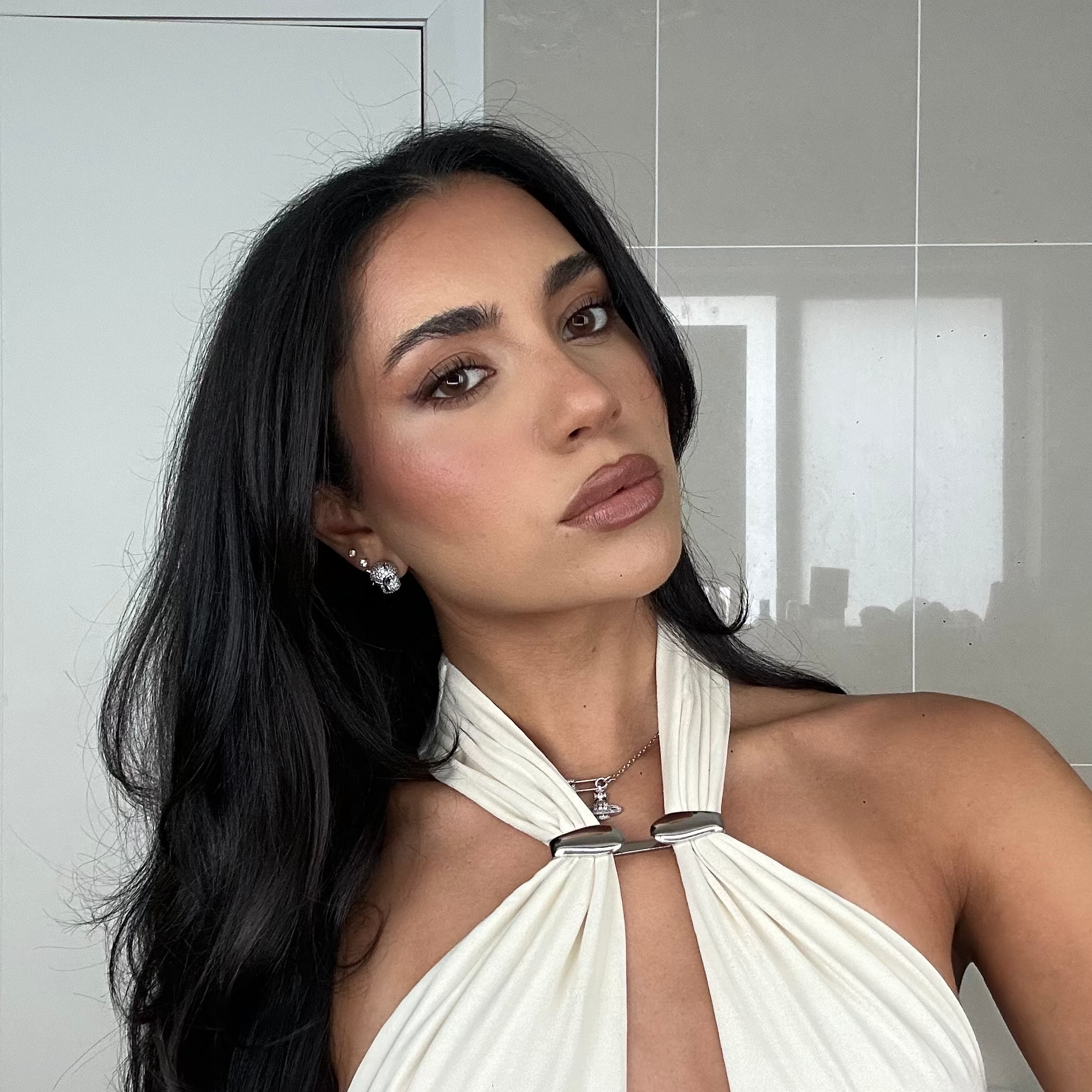 A woman with long black hair wearing a cream halter-neck dress with a front keyhole cutout and silver-toned metal hardware, posing in front of a light gray tiled wall.